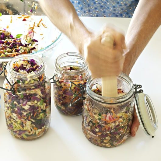 Three glass jars filled with Sauerkraut are on a table. A pair of hands pound a pestle into the far right jar.