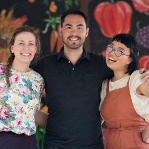 Joyce Liao, Meryn Corkery and Colin Dring stand smiling and arm-in-arm in front of a painted mural of vegetables.