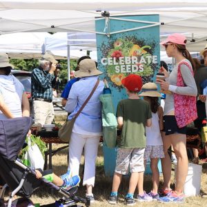 Three people standing at a booth under a tent. A sign reads "celebrate seeds."