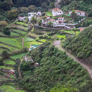 A photo from above a hillside on Tenerife showing a terraced farm with several small fields at different levels cut into the hillside, several buildings and houses at the top of the fields, and a road cut into the hillside above the farm.