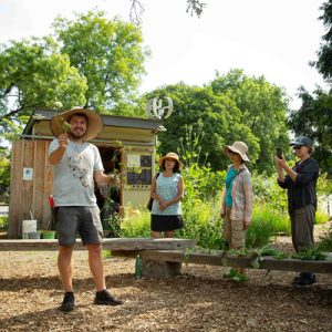 A smiling man in a straw hat showing flowers to a group in a community garden